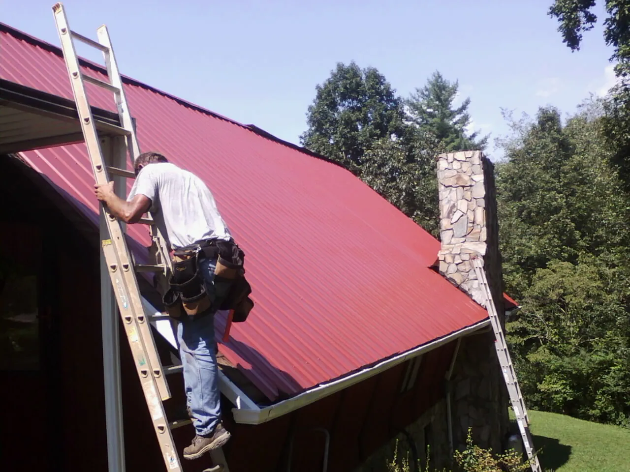 Roofer on a ladder beside a red metal roof and stone chimney — Soffit & Fascia Repair by Roofing Brothers in Hartford City